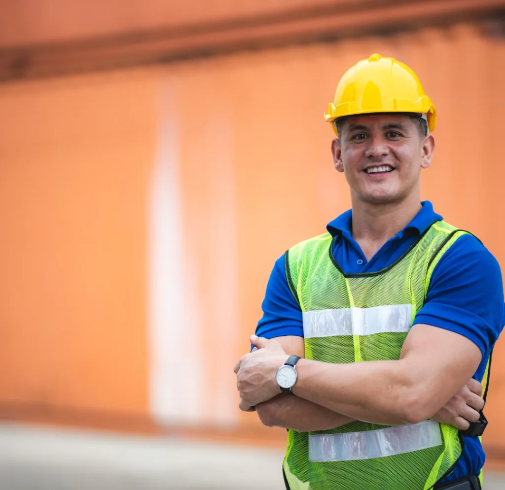 Professional affordable residential builder -
a smiling construction worker wearing a yellow hard hat and reflective safety vest stands confidently with arms crossed in front of an orange building.