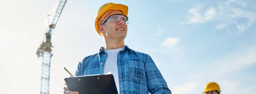Affordable residential builders - a construction worker wearing a yellow hard hat and protective glasses stands outdoors holding a clipboard. A construction crane is visible in the background under a bright blue sky.