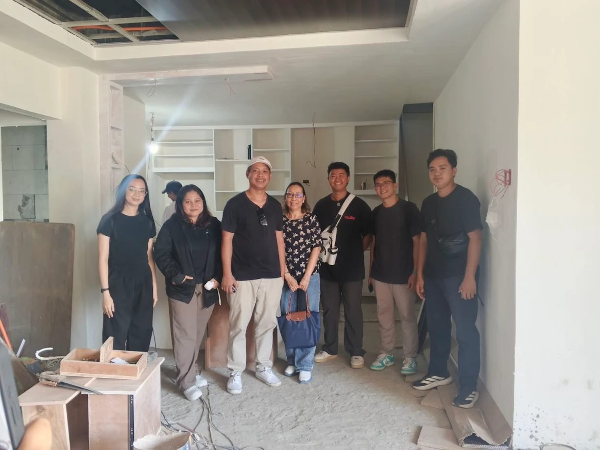 A group of seven people smiling for a photo inside a building under construction, featuring white walls and unfinished shelving.