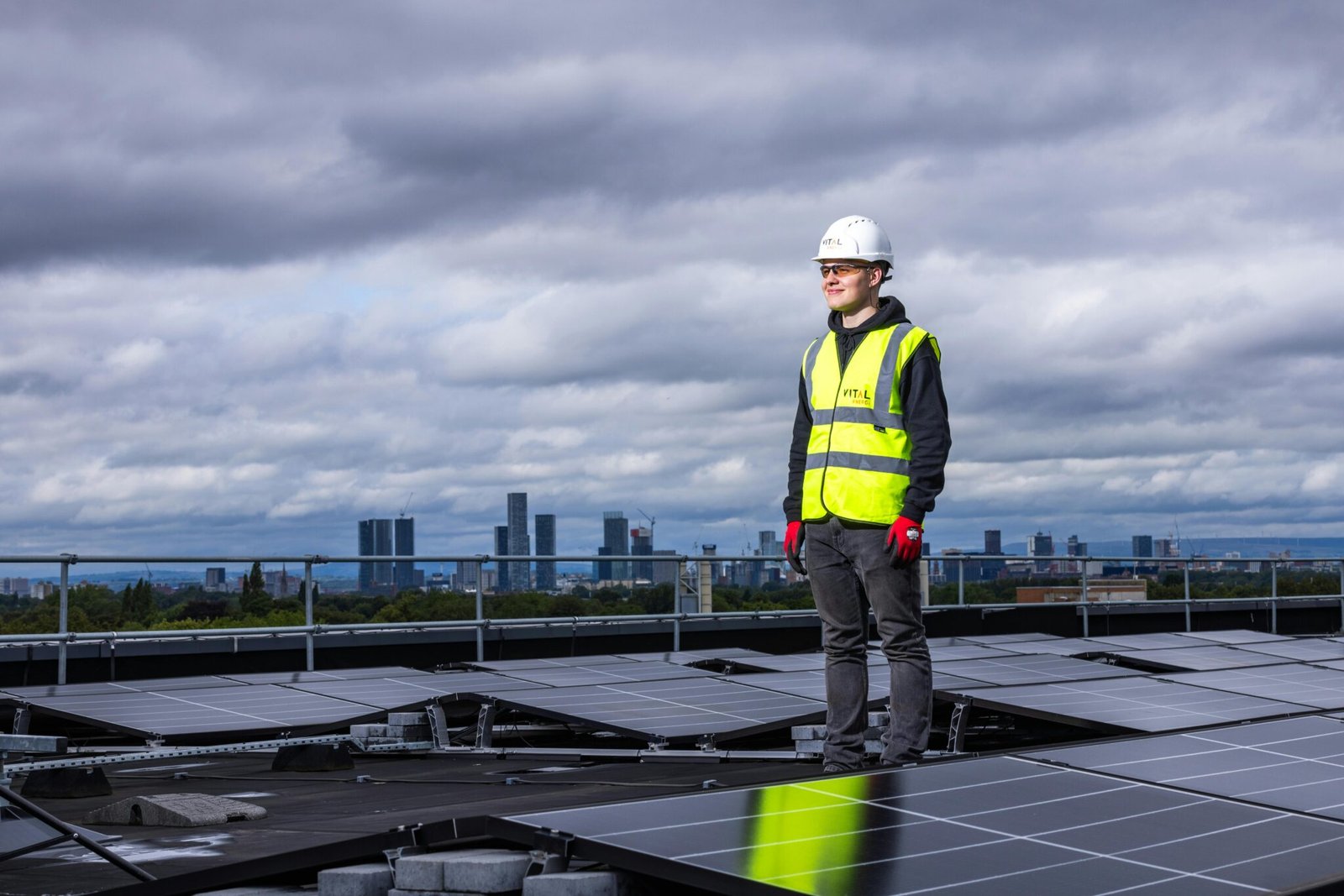 Sustainable construction with an engineer standing on rooftop inspecting solar panels with city skyline in background.